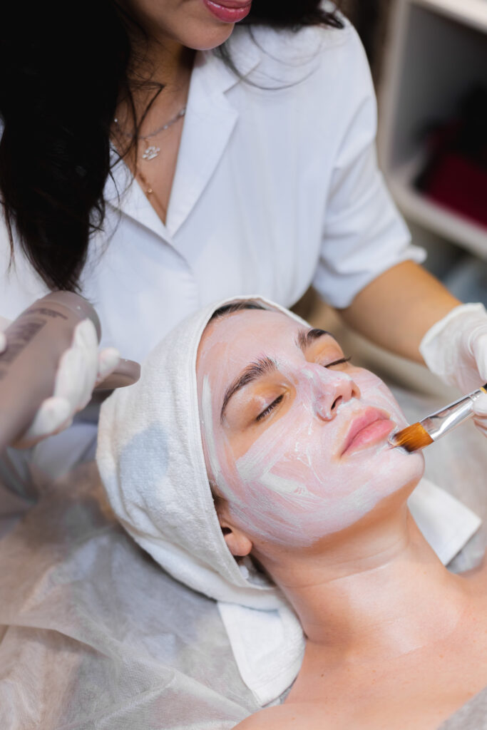 Una esteticista aplica con una brocha una mascarilla hidratante blanca en el rostro de una joven clienta en un salón de belleza y spa. Una esteticista aplica con una brocha una mascarilla hidratante blanca en el rostro de una joven clienta en un salón de belleza y spa.